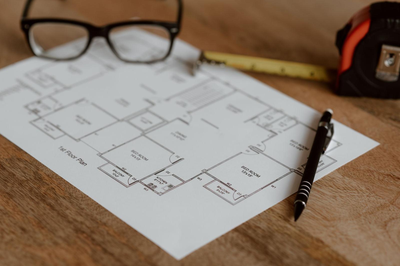 A house floor plan on a wooden desk with a pen, a tape measure and a pair of glasses
